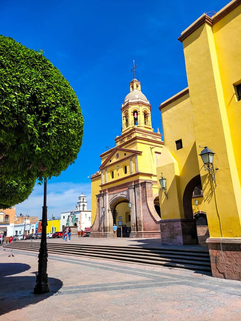 A view of a beautiful yellow church in Queretaro with lush green trees and bright blue skies.
