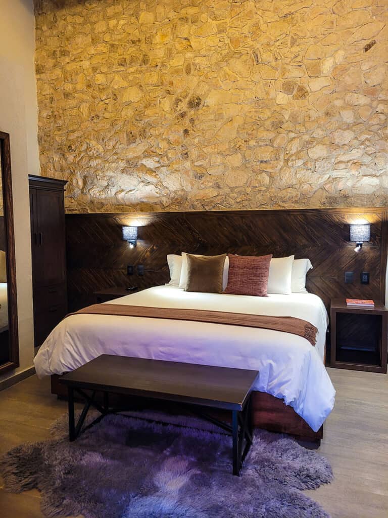 A room at Casa Once hotel in Queretaro with a natural stone wall, warm wood accents and a fluffy inviting king bed.