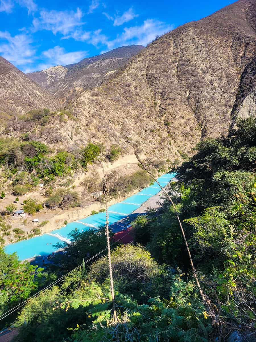 The blue Tolantongo river cuts through the box canyon with mountains and a blue sky behind it.