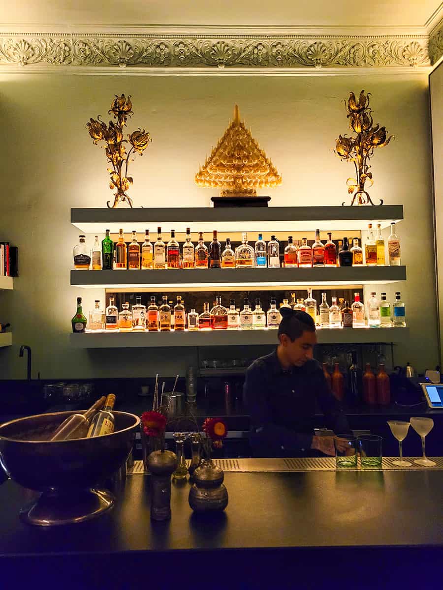 A bartender mixing a drink at the bar of Salon Rosetta with liquor bottles lit up behind him.