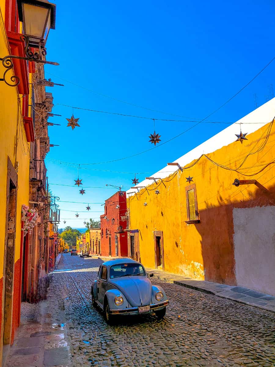 A blue old fashioned VW Beetle drives down orange and yellow colonial streets in San Miguel de Allende.